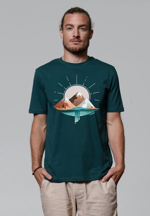 Teal cotton t-shirt featuring a graphic of mountains and sun, with warm earthy colors and a water element at the base. Short sleeves.