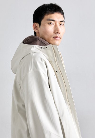 Chaqueta impermeable blanca con capucha, con un forro interior marrón, cierre con cremallera y sutiles detalles en negro. Textura suave y diseño minimalista.