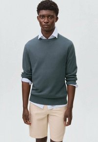 Dark green sweater with crew neck, layered over light blue collared shirt. Worn with light beige shorts. Smooth fabric, casual fit.