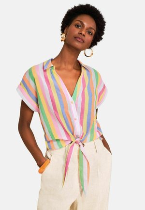 Kobieta ubrana w krótki rękaw, wielokolorową pionową koszulę zapinaną na guziki, zawiązaną w talii, beżowe spodnie, duże kolczyki-koła i pomarańczową bransoletkę.