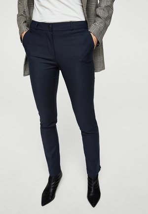 Persona vistiendo pantalones ajustados azul marino, botas negras puntiagudas, camiseta blanca y blazer gris con cuadros, con las manos en los bolsillos.