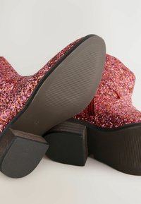 Bottines à paillettes rouges et roses avec un talon en bois épais et une semelle en caoutchouc. Dotées d'une surface texturée et d'un détail de fermeture éclair sur le côté.
