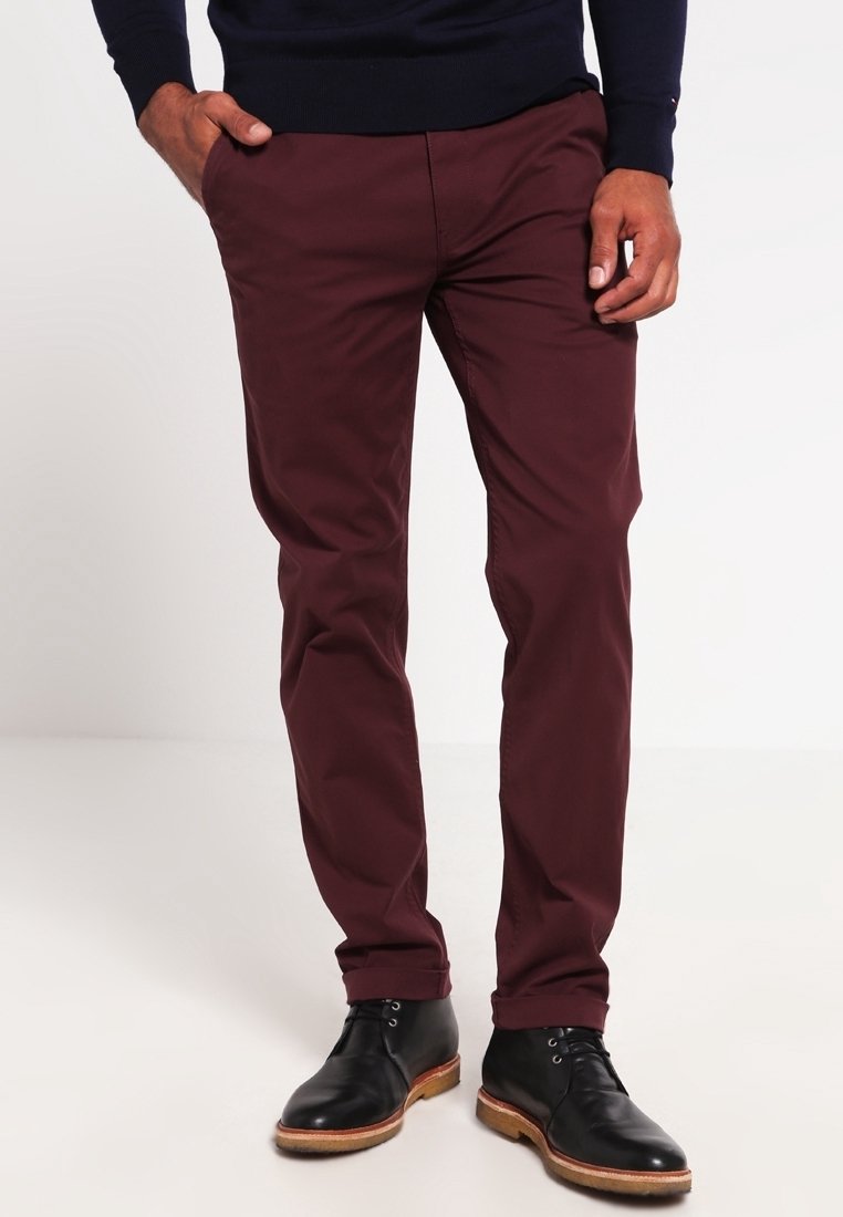 Homme portant un pantalon slim bordeaux foncé, des bottines en cuir noires et un haut à manches longues foncé, les mains dans les poches.
