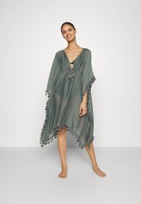 Kaftano verde trasparente con scollatura a V, vita con cordoncino e bordi con frange, caratterizzato da una forma asimmetrica e tessuto leggero.