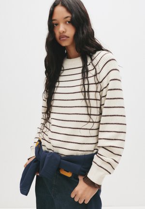 Maglione lavorato a maglia color crema con righe marrone scuro, caratterizzato da un collo rotondo e polsini a costine. Felpa blu legata in vita.
