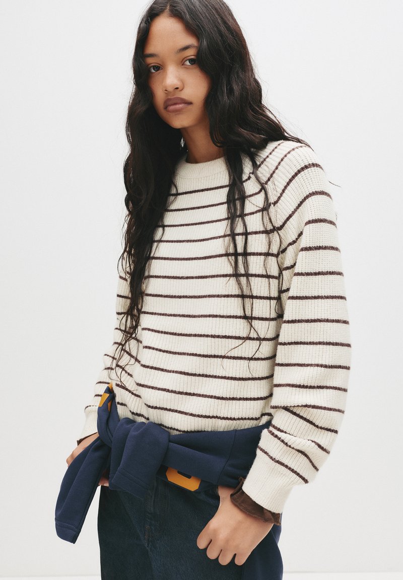 Strickpullover in Creme mit dunkelbraunen Streifen, runder Ausschnitt und gerippte Bündchen. Blauer Sweatshirt um die Taille gebunden.