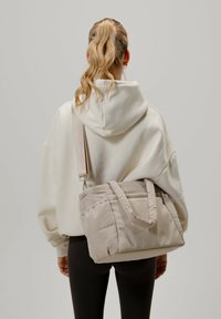 Sac à épaule beige rembourré avec poignées doubles, texture douce et bandoulière réglable, associé à un sweat à capuche clair et des leggings.