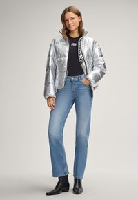 Silberne Pufferjacke mit glänzender Oberfläche, Reißverschluss und hohem Kragen. Getragen über einem schwarzen Shirt, kombiniert mit hellblauen Jeans und schwarzen Ankle Boots.