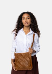 Donna con capelli ricci che indossa una camicia bianca e una gonna marrone, mentre tiene una borsa a tracolla in pelle intrecciata marrone su uno sfondo neutro.