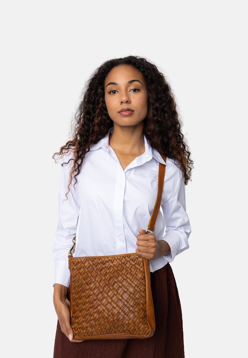 Donna con capelli ricci che indossa una camicia bianca e una gonna marrone, mentre tiene una borsa a tracolla in pelle intrecciata marrone su uno sfondo neutro.