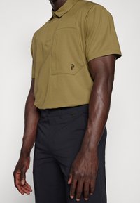 Oliwkowa koszulka polo z krótkim rękawem, z kieszenią na piersi i subtelnym logo; w połączeniu z ciemnoniebieskimi spodniami, o gładkiej teksturze i dopasowanym kroju.