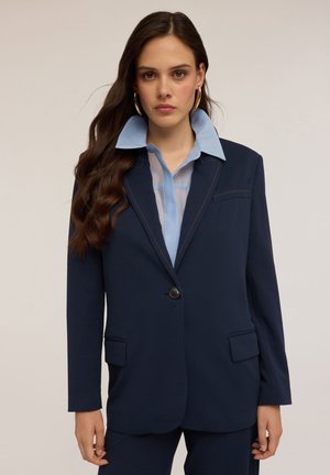 Blazer blu navy con rever a punta e chiusura a un bottone. Presenta una camicia azzurra sotto e tasche frontali. Tessuto morbido con cuciture a contrasto.