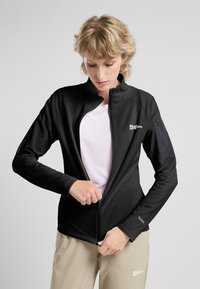 Zwarte zip-up jack met een gestructureerde afwerking, hoge kraag en een zijzak. Gedragen over een lichtroze basislaag en beige broek.