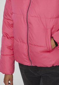 Jachetă pufoasă roz aprins, cu închidere prin fermoar, textură ușor umflată și buzunare laterale. Dispune de mâneci lungi, largi și o silhouette casuală.