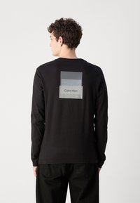Melna garām piedurknēm krekls ar taisnstūrveida grafiku uz muguras, kurā ir dažādi pelēko toņu nokrāsas un uzraksts "Calvin Klein." Kokvilnas materiāls.