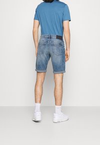 Denim shorts in light blue with rolled cuffs, featuring back pockets and a dark label on the waistband. Paired with a blue t-shirt and white sneakers.