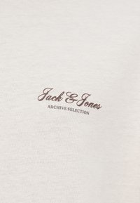 Ανοιχτό μπεζ βαμβακερό μπλουζάκι με καφέ λογότυπο "Jack & Jones" και την επιγραφή "ARCHIVE SELECTION" σε καμπυλωτό κείμενο από κάτω.