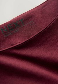 Burgundy cotton fabric of a shirt collar, featuring a smooth texture and a printed label with "NEXT" in dark green.