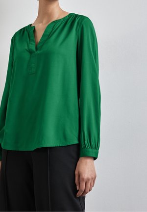 Femme portant un chemisier vert uni à manches longues avec un col en V et un pantalon noir ample devant un mur clair uni.