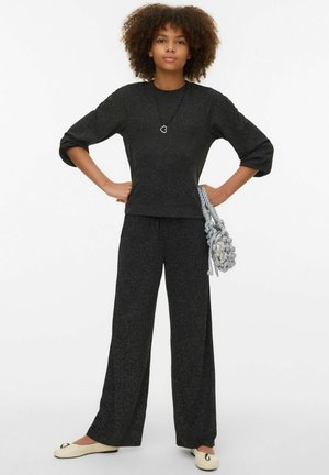 Zwarte gebreide trui met driekwart mouwen gecombineerd met bijpassende wijde broekspijpen. Geaccessoriseerd met een gevlochten grijze tas en een hartvormige ketting.