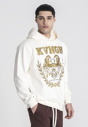 Hoodie krem boje s prednjim grafičkim prikazom u zlatnoj boji koji prikazuje lava i dekorativni tekst. Tkanina izgleda mekano; ima veliki prednji džep i kapuljaču s vezicom.