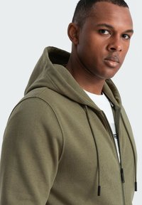 Olivgrüne Kapuzenjacke aus Baumwollmischung, mit Reißverschluss vorne, verstellbarem Kordelzug an der Kapuze, gerippten Bündchen und glatter Textur.
