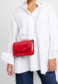 Rød skuldertaske i laklæder med klaplukning og sølvkæde, vist mod en hvid skjorte. Har en glat, skinnende overflade.