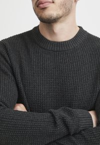 Sweter dzianinowy w ciemnoszarym kolorze, z prążkowanym dekoltem i mankietami, teksturowanym splotem oraz luźnym krojem. Zbliżenie ukazuje subtelne wzory.