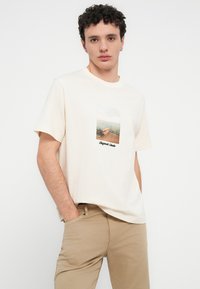 Κρεμ βαμβακερό t-shirt με κοντά μανίκια, με εκτυπωμένη γραφική απεικόνιση μιας παραθαλάσσιας θέας και το κείμενο "Alguien's Studio" στο κάτω μέρος.