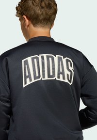 Svart sportjacka med stor vit "ADIDAS"-text i fet stil på baksidan, med en slät, glänsande yta och ribbat krage.