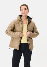 Lichtbruin geïsoleerd jack met een capuchon, met een geborduurd ontwerp en grijze binnenvoering, gecombineerd met een gele top en donkere broek.