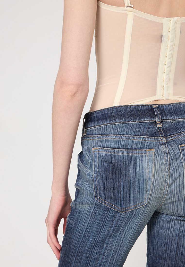 Beige-Mesh-Bodysuit mit verstellbaren Trägern, kombiniert mit blauen, gestreiften Denim-Jeans mit zwei Gesäßtaschen und einem leicht ausgefransten Saum.