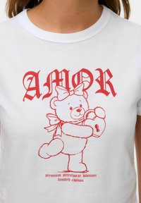 Hvid bomulds t-shirt med et rødt grafisk tryk af en bjørn, der holder en hjerteformet lås, tekst "AMOR" og "premium streetwear division limited edition."