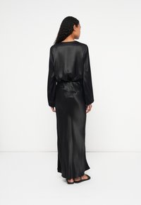 Rochie maxi din satinat negru, cu mâneci lungi, o croială relaxată și talie strânsă. Prezintă o textură netedă și un decolteu rotund simplu.