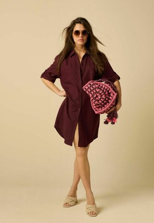 Femme aux longs cheveux portant une robe chemise bordeaux et des sandales beiges, tenant un sac crocheté rose et marron, portant des lunettes de soleil rondes sur fond beige.