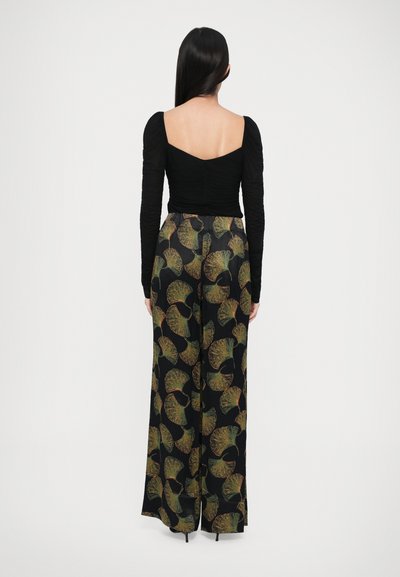 Top negru, cu mâneci lungi și textură pliată pe spate, asortat cu pantaloni largi cu un model de frunze în verde și aur pe un fundal negru.