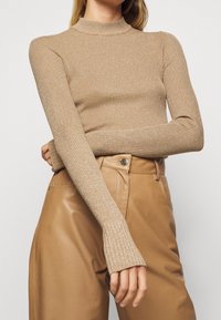 Femme portant un pull col roulé beige côtelé ajusté et un pantalon en cuir brun taille haute, bras croisés à la taille, sur un fond uni.