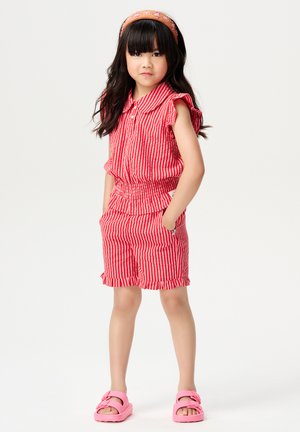 Ensemble sans manches à rayures rouges et blanches avec des épaules volantées, taille élastique et shorts ; associé à des sandales roses et à un bandeau floral.