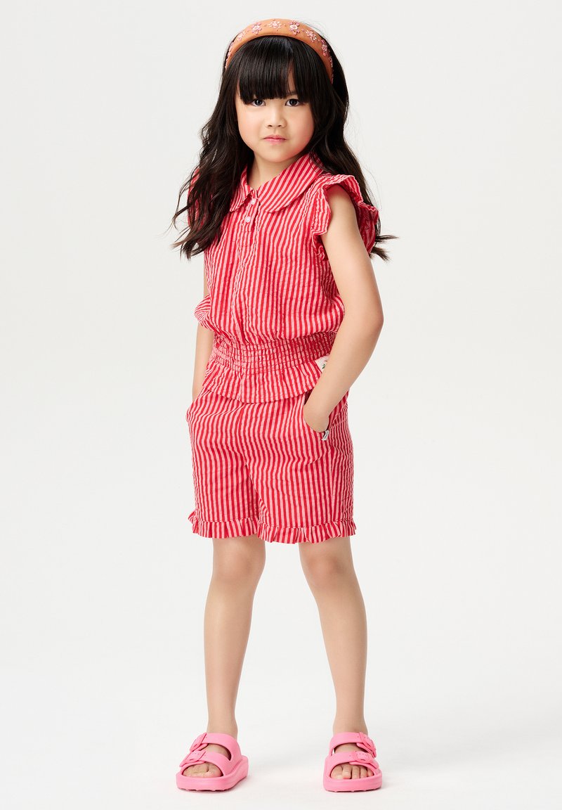 Ensemble sans manches à rayures rouges et blanches avec des épaules volantées, taille élastique et shorts ; associé à des sandales roses et à un bandeau floral.