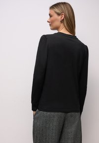 Schwarzes Langarmshirt mit rundem Ausschnitt und gepufften Schultern, kombiniert mit grauen, strukturierten Hosen. Glatter Stoff, figurbetonter Schnitt.