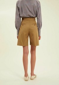 Shorts marron cintrés en tissu lisse, avec une taille haute, deux poches arrière avec des boutons, et une coupe droite.