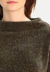 Haut en velours côtelé vert olive avec un col large, présentant une texture à côtes verticales et un tissu doux et moelleux. Aucune quincaillerie ou motif visible.