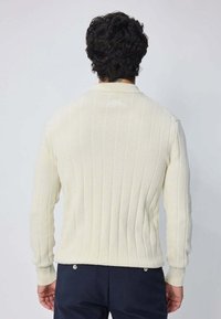 Homme avec des cheveux bouclés noirs portant un pull crème à manches longues en côte et un pantalon sombre, se tenant de dos contre un fond uni.