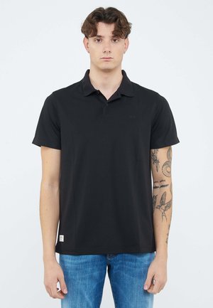 Giovane uomo con capelli ricci che indossa una polo nera e jeans blu, mostrando tatuaggi dettagliati sul braccio destro.