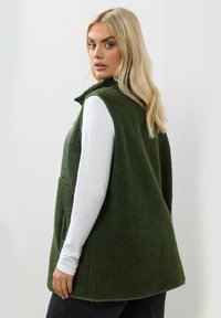 Grön fleeceregler med en mjuk, texturerad yta och dragkedja framtill. Har breda ärmhål och sidofickor. Bärs över en vit långärmad tröja.