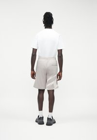 Moški s črnimi pletenicami stoji obrnjeno stran, oblečen v belo majico, svetlo sive kratke hlače z vzorcem, bele nogavice in črne superge.