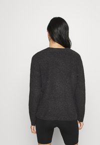 Maglione grigio scuro lavorato a maglia con scollo rotondo e vestibilità ampia, caratterizzato da una superficie strutturata. Indossato sopra pantaloncini neri.