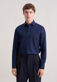 Camicia a maniche lunghe color blu navy realizzata in tessuto liscio, con colletto classico e chiusura frontale con bottoni. Abbinata a pantaloni scuri.