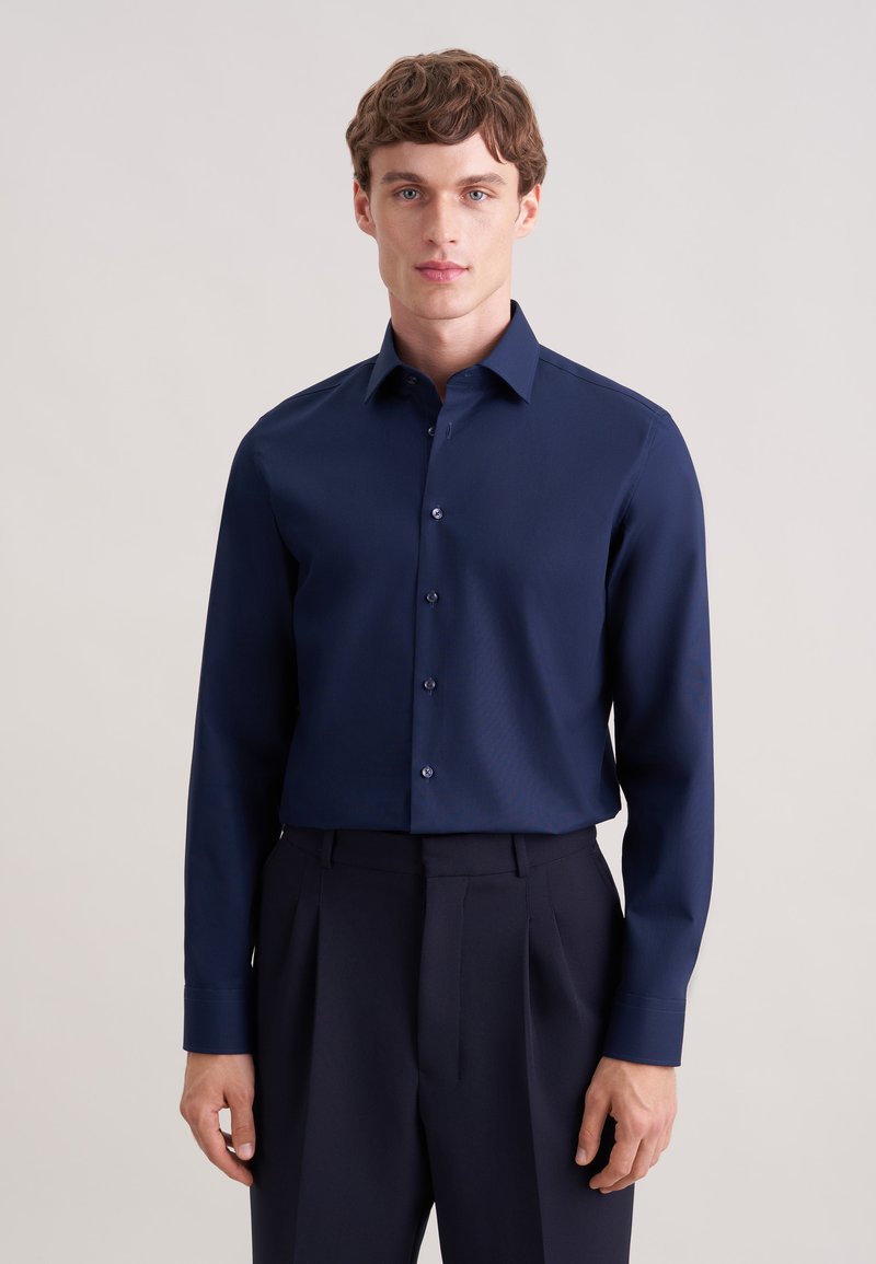 Camicia a maniche lunghe color blu navy realizzata in tessuto liscio, con colletto classico e chiusura frontale con bottoni. Abbinata a pantaloni scuri.