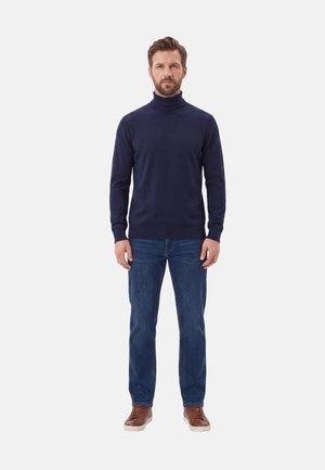 Uomo in piedi di fronte, indossa un maglione dolcevita blu navy, jeans blu e sneaker in pelle marrone su sfondo bianco.